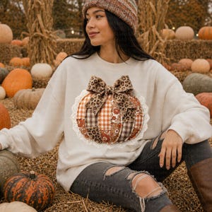 May include: Cream-colored sweatshirt featuring a patchwork pumpkin design with a leopard print bow. The pumpkin is made of various fabrics in shades of brown, orange, and white, and is trimmed with white lace. The sweatshirt is a fall-themed item.
