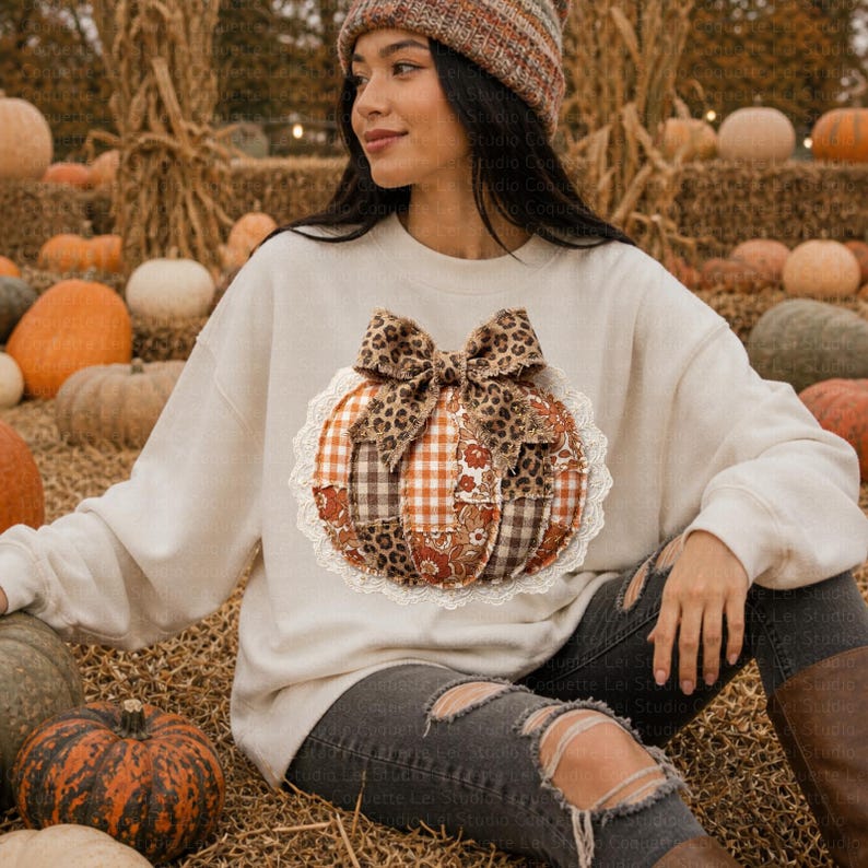 May include: Cream-colored sweatshirt featuring a patchwork pumpkin design with a leopard print bow. The pumpkin is made of various fabrics in shades of brown, orange, and white, and is trimmed with white lace. The sweatshirt is a fall-themed item.