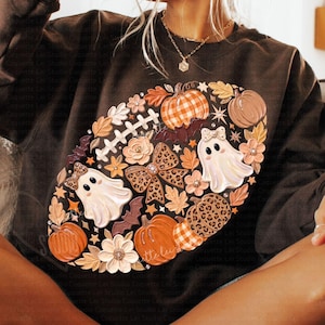 Op de afbeelding: Een donkerbruin sweatshirt met een grote ovale afbeelding in de vorm van een football, gevuld met pompoenen, spookjes met strikjes, strikjes met luipaardprint, herfstbladeren, bloemen en vleermuizen in oranje, witte en bruine tinten.