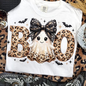 May include: A white t-shirt featuring the text BOO in a leopard print pattern. The letter O is replaced by a ghost wearing a large black lace bow. The design includes small black bats, gold stars, and scattered rhinestones across the graphic.