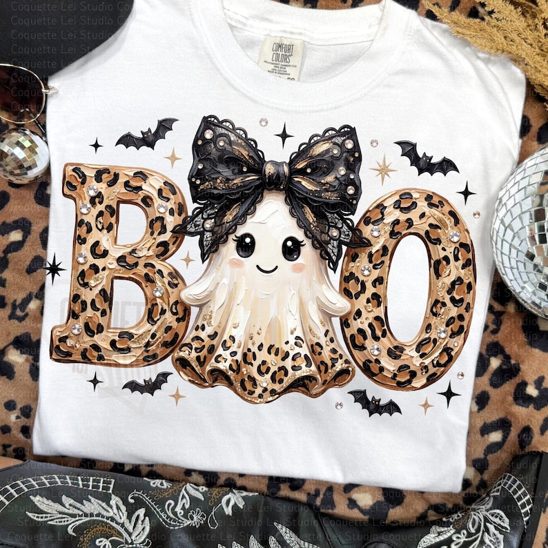 May include: A white t-shirt featuring the text BOO in a leopard print pattern. The letter O is replaced by a ghost wearing a large black lace bow. The design includes small black bats, gold stars, and scattered rhinestones across the graphic.
