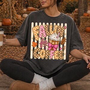 May include: Dark grey t-shirt featuring the text "Fallin' for Coffee" in a checkered and pink design. The graphic includes a coffee cup with whipped cream, a pink bow, and autumn leaves. The shirt has a relaxed fit.