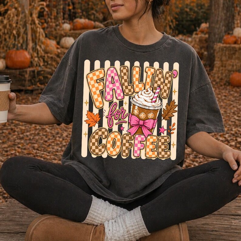 May include: Dark grey t-shirt featuring the text "Fallin' for Coffee" in a checkered and pink design. The graphic includes a coffee cup with whipped cream, a pink bow, and autumn leaves. The shirt has a relaxed fit.
