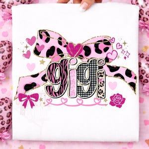 K&ouml;nnte beinhalten: Wei&szlig;es T-Shirt mit dem Namen "GIGI" in einem dekorativen Design. Die Buchstaben haben Zebra-, Hahnentritt- und Leopardenmuster. Eine rosa Schleife mit einem schwarz-rosa Kuhdruck, Herzen und eine Rose vervollst&auml;ndigen das Design.