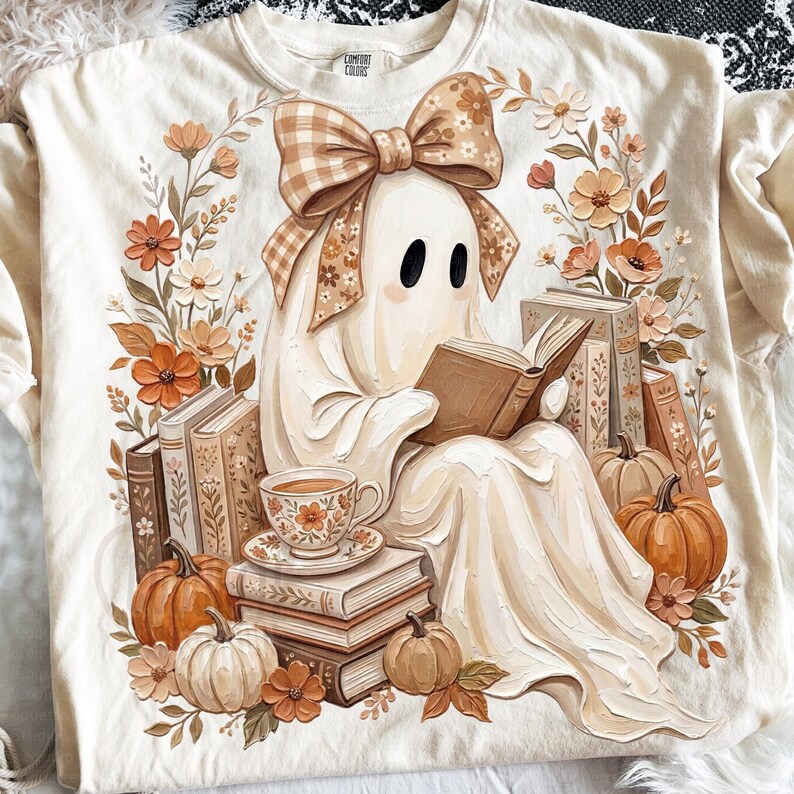 Puede incluir: Una camiseta de color crema con un gr&aacute;fico de un fantasma que lleva un gran lazo de cuadros y lee un libro. El fantasma est&aacute; rodeado de pilas de libros, calabazas naranjas y elementos florales en tonos marrones, naranjas y crema.