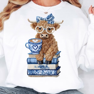 May include: White sweatshirt featuring a watercolor illustration of a Highland cow wearing heart-shaped glasses and a blue bow. The cow is holding a blue and white teacup with a heart design and is sitting on a stack of blue books.
