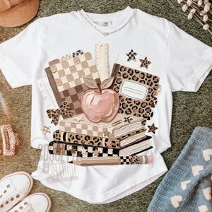 May include: White t-shirt featuring a stack of books, an apple, and a pencil. The books have a checkered pattern, leopard print, and star accents. A ruler and a bow are also included in the design. The text "2020 Lei Studio" is printed on the shirt.