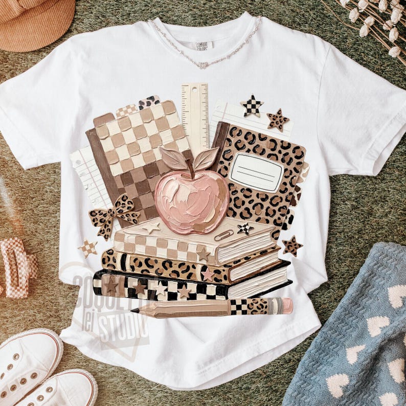 May include: White t-shirt featuring a stack of books, an apple, and a pencil. The books have a checkered pattern, leopard print, and star accents. A ruler and a bow are also included in the design. The text "2020 Lei Studio" is printed on the shirt.