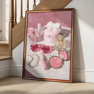 May include: A framed artwork featuring a collection of pink accessories. The composition includes heart-shaped sunglasses, a pearl necklace, a compact mirror, perfume bottles, and a heart-shaped box with a bow. The color palette is soft, with pink and white tones.