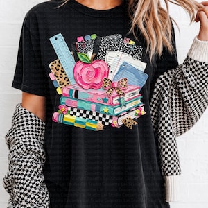 May include: Black t-shirt featuring a colorful graphic of books, an apple, and school supplies. The design includes a stack of books with a pink apple, a ruler, and notebooks. The graphic has a back-to-school theme.