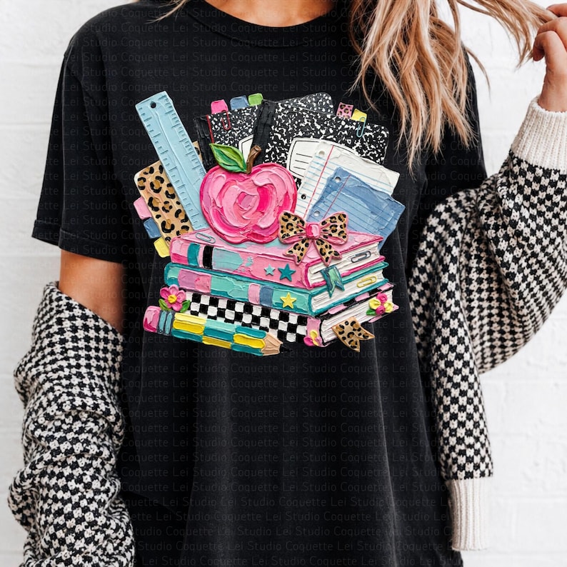 May include: Black t-shirt featuring a colorful graphic of books, an apple, and school supplies. The design includes a stack of books with a pink apple, a ruler, and notebooks. The graphic has a back-to-school theme.