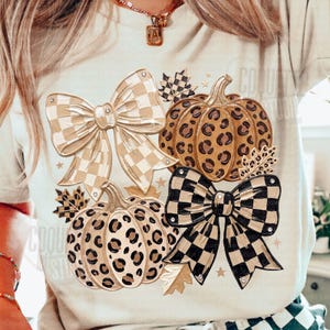 May include: A cream-colored t-shirt featuring a graphic print of two pumpkins with leopard print patterns, two large bows with checkered patterns in beige and black, and several small autumn leaves and star shapes scattered around the design.