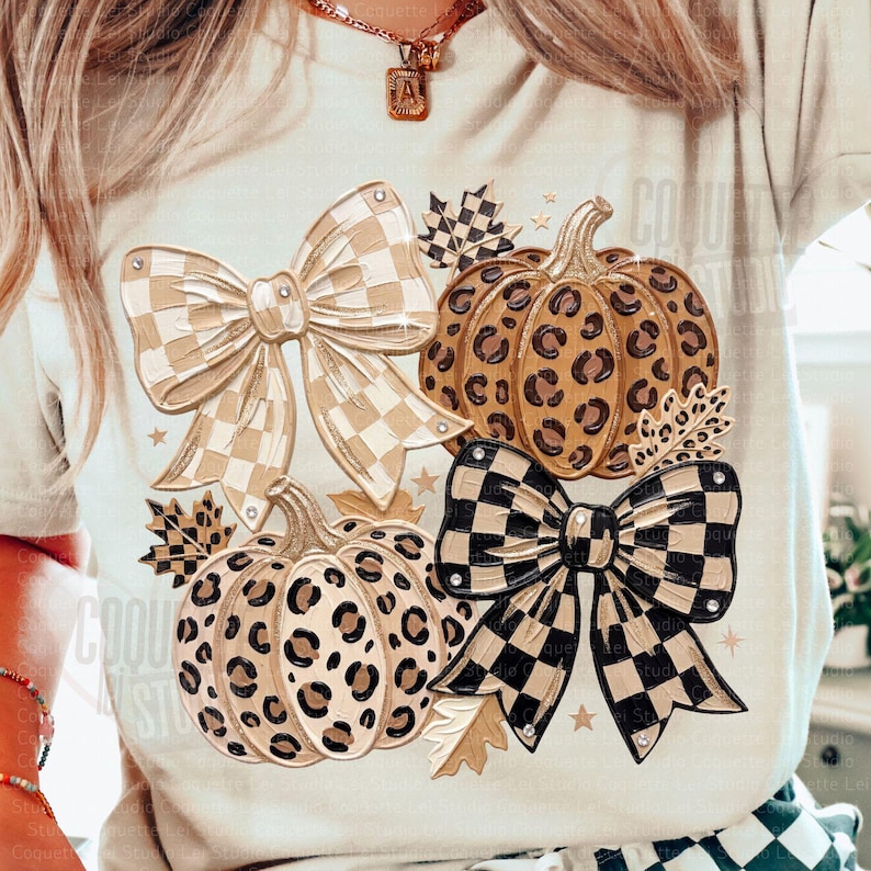 May include: A cream-colored t-shirt featuring a graphic print of two pumpkins with leopard print patterns, two large bows with checkered patterns in beige and black, and several small autumn leaves and star shapes scattered around the design.