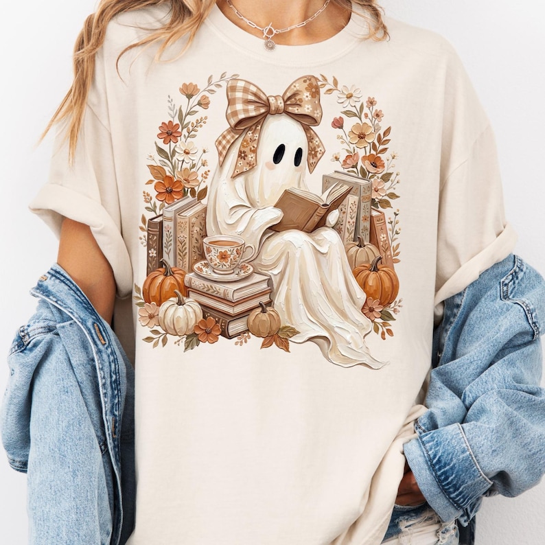 Puede incluir: Una camiseta de color crema con un gr&aacute;fico de un fantasma que lleva un lazo de cuadros marr&oacute;n, leyendo un libro y rodeado de pilas de libros, calabazas naranjas, una taza de t&eacute; y flores de oto&ntilde;o en tonos marrones, naranjas y crema.