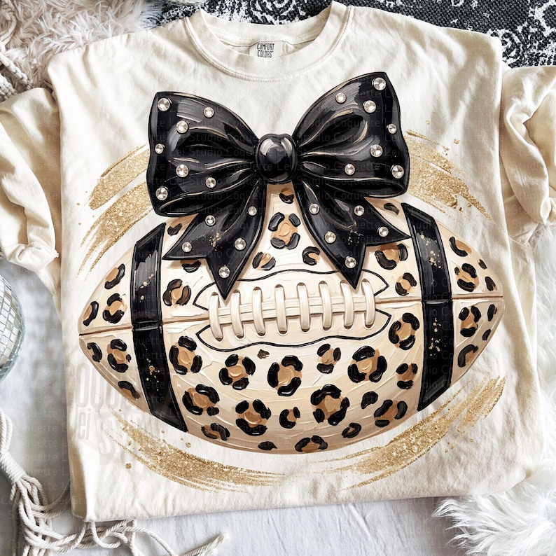 May include: A cream-colored t-shirt featuring a graphic of a football with a leopard print pattern, black stripes, and a large black bow adorned with clear rhinestones. The design is centered on the shirt and accented by a gold glitter brushstroke.