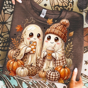 May include: A dark brown sweatshirt featuring a graphic of two ghosts. One ghost wears a leopard print pattern and holds a pumpkin-themed phone. The other ghost wears a brown knit beanie and holds a coffee cup. Surrounded by orange pumpkins and leaves.