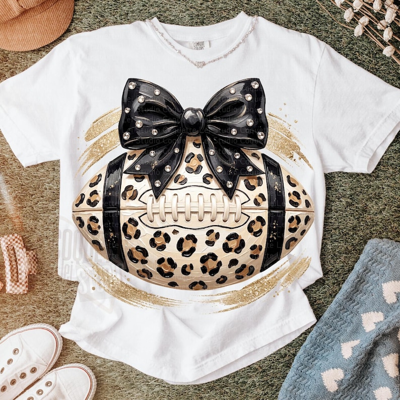May include: A white short-sleeve t-shirt featuring a graphic of a football with a tan and black leopard print pattern. A large black bow with silver stud accents sits on top of the football. The design is framed by gold glitter brush strokes.