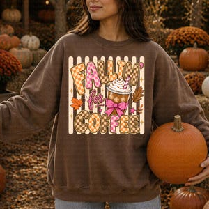 May include: A brown sweatshirt featuring the phrase "Fallin' for Coffee" in a checkered pattern. The design includes a coffee cup with whipped cream, a pink bow, and autumn leaves. This is a fall-themed item of clothing.