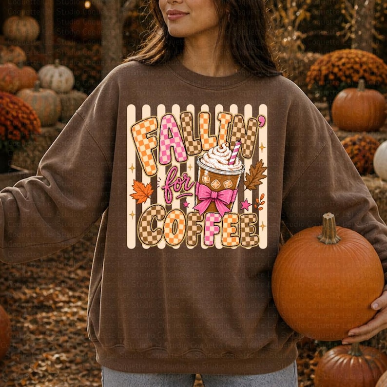 May include: A brown sweatshirt featuring the phrase "Fallin' for Coffee" in a checkered pattern. The design includes a coffee cup with whipped cream, a pink bow, and autumn leaves. This is a fall-themed item of clothing.
