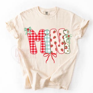 May include: Cream-colored t-shirt with the word "MINI" in a patchwork design. The letters feature red gingham, mint green plaid, and strawberry patterns, accented with bows and scalloped edges. A cute and playful design.