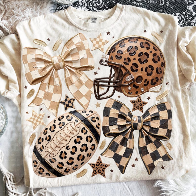 May include: A cream-colored t-shirt featuring a graphic print of a leopard print football helmet, a leopard print football, a beige checkered bow, a black and beige checkered bow, and several small stars scattered across the front fabric.