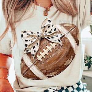 May include: A beige t-shirt featuring a graphic of a brown football with a large white bow decorated with black spots. The football has a textured, painted appearance with white laces and white stripes on the ends, centered on the front of the shirt.
