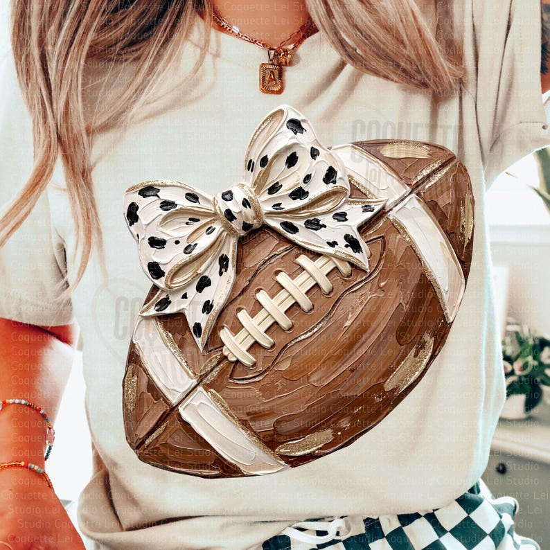 May include: A beige t-shirt featuring a graphic of a brown football with a large white bow decorated with black spots. The football has a textured, painted appearance with white laces and white stripes on the ends, centered on the front of the shirt.