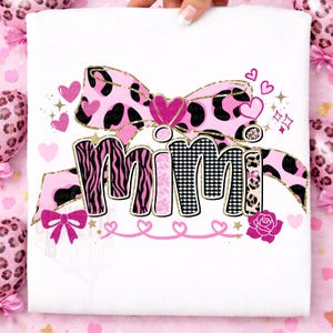 K&ouml;nnte beinhalten: Wei&szlig;es T-Shirt mit dem Wort "MIMI" in einem verspielten Design. Die Buchstaben sind mit Zebrastreifen, rosa Mustern und einem schwarz-wei&szlig;en Hahnentrittmuster verziert. Eine rosa Schleife und Herzen vervollst&auml;ndigen das Design.