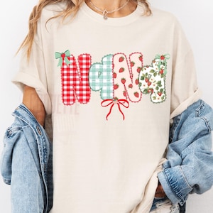 May include: Beige t-shirt with the word "NANA" in a colorful, patchwork design. The letters feature red gingham, green plaid, strawberries, and floral patterns, accented with bows. A casual, comfortable top.