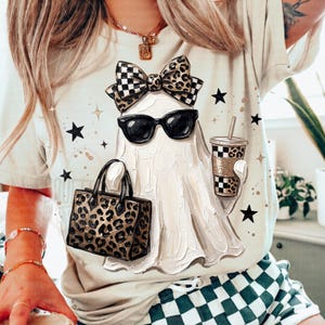 Può includere: Una t-shirt color crema con la grafica di un fantasma che indossa occhiali da sole neri e un fiocco con motivo leopardato e a scacchi. Il fantasma tiene in mano una tazza e una borsa con motivo leopardato e a scacchi, circondato da stelle nere.