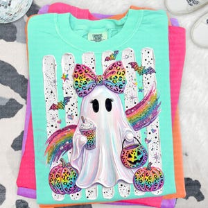 May include: A mint green t-shirt featuring a graphic of a white ghost with a large leopard print bow, holding a leopard print coffee cup and a jack-o'-lantern bucket. The design includes rainbow wings, leopard print pumpkins, and bats.