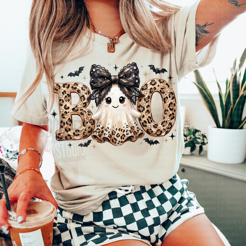 May include: A beige short-sleeve t-shirt featuring a graphic of a ghost with a black lace bow. The word BOO is printed in large leopard print letters, with the ghost replacing the middle letter. Small black bats and stars surround the design.