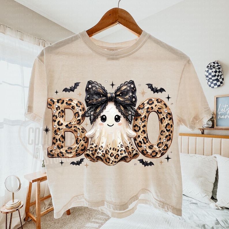 May include: A beige short-sleeve t-shirt featuring a graphic of a ghost with a large black lace bow on its head. The ghost is flanked by the letters B and O, both filled with a leopard print pattern. Small black bats and stars surround the design.