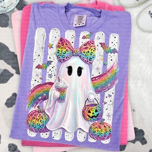 May include: A purple short-sleeve t-shirt featuring a graphic of a white ghost wearing a rainbow leopard print bow. The ghost holds a coffee cup and a jack-o'-lantern bucket, surrounded by rainbow leopard pumpkins, bats, and stars.