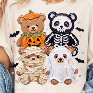 May include: A cream-coloured t-shirt featuring a graphic of four teddy bears dressed for Halloween. One bear wears a pumpkin costume, one is a skeleton, one is wrapped in mummy bandages, and one is dressed as a ghost, surrounded by small bats.