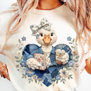 Może przedstawiać: Biała koszulka z postacią kaczki w dżinsowej kurtce, trzymającej książkę i kawę. Kaczka ma kwiatową kokardę i jest otoczona niebieskimi i kremowymi kwiatami, tworząc fantazyjny wzór w kształcie serca.