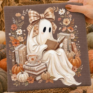 May include: A square fabric panel featuring a white ghost wearing a tan gingham bow, reading a book while seated among stacks of books, pumpkins, and a floral teacup. The illustration is framed by a wreath of autumn-colored flowers on a brown background.