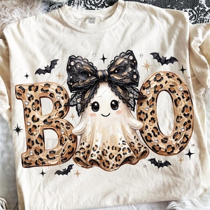 May include: A cream-colored t-shirt featuring the text BOO in a brown and black leopard print pattern. A central ghost illustration wears a large black lace bow. The design includes small black bats, gold star accents, and clear rhinestones.