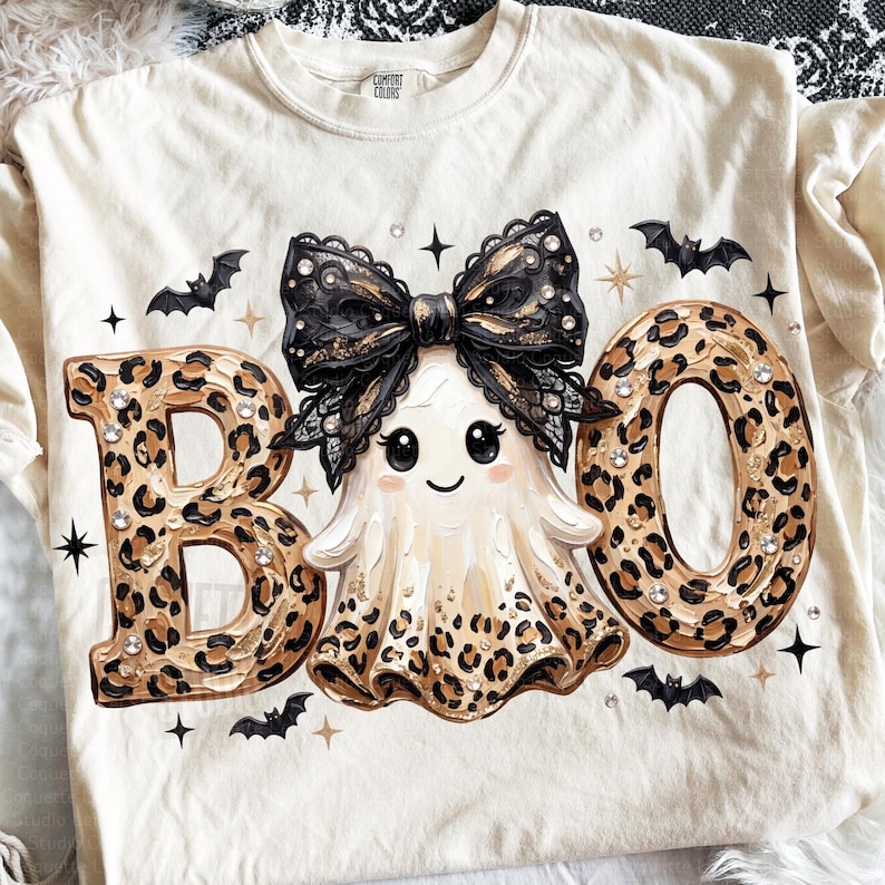 May include: A cream-colored t-shirt featuring the text BOO in a brown and black leopard print pattern. A central ghost illustration wears a large black lace bow. The design includes small black bats, gold star accents, and clear rhinestones.