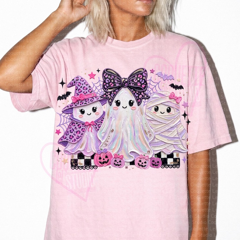 May include: A light pink t-shirt featuring a graphic of three cartoon ghosts. One ghost wears a purple leopard print witch hat, the center ghost has a large black leopard print bow, and the third is wrapped like a mummy. Small pumpkins and bats surround them.