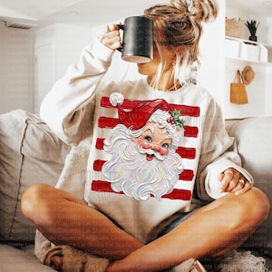 K&ouml;nnte beinhalten: Ein cremefarbenes Langarm-Sweatshirt mit einem Grafikdruck eines weihnachtlichen Santa-Claus-Gesichts im Vintage-Stil, mit roter M&uuml;tze und wei&szlig;em Bart, &uuml;berlagert von horizontalen roten Streifen. Das Sweatshirt hat einen Rundhalsausschnitt.