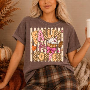May include: A dark grey t-shirt featuring the phrase "Fallin' for Coffee" in a colourful, chequered design. The graphic includes a coffee cup with whipped cream, a pink bow, and autumn leaves. The shirt is worn with plaid trousers.