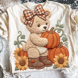 May include: A cream-coloured t-shirt featuring a textured graphic of a tan teddy bear wearing a brown and white checkered bow. The bear holds an orange pumpkin, surrounded by additional pumpkins, green vines, and yellow sunflowers.