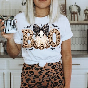 May include: A white short-sleeved t-shirt featuring a graphic of a ghost with a black lace bow, surrounded by the word BOO in leopard print lettering. The design includes small black bats and stars. The shirt is paired with leopard print leggings.