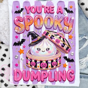 May include: A white t-shirt featuring a graphic of a cute dumpling in a bamboo steamer wearing a witch hat. The text reads YOU'RE A SPOOKY DUMPLING in pink and glittery orange letters. The design includes purple brush strokes, bats, and stars.