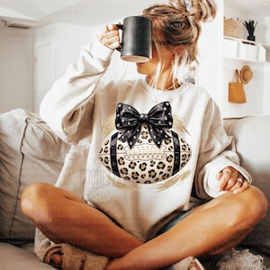 May include: A cream-colored long-sleeve sweatshirt featuring a central graphic of an American football with a leopard print pattern, accented by a large black bow with white dots and gold brushstroke details on the front of the garment.