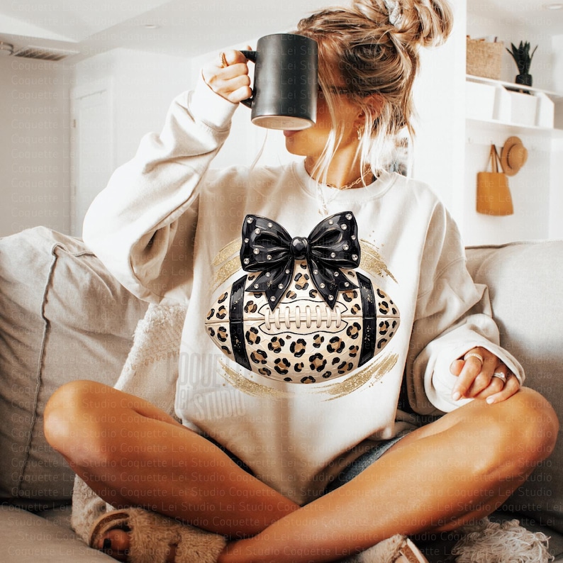 May include: A cream-colored long-sleeve sweatshirt featuring a central graphic of an American football with a leopard print pattern, accented by a large black bow with white dots and gold brushstroke details on the front of the garment.