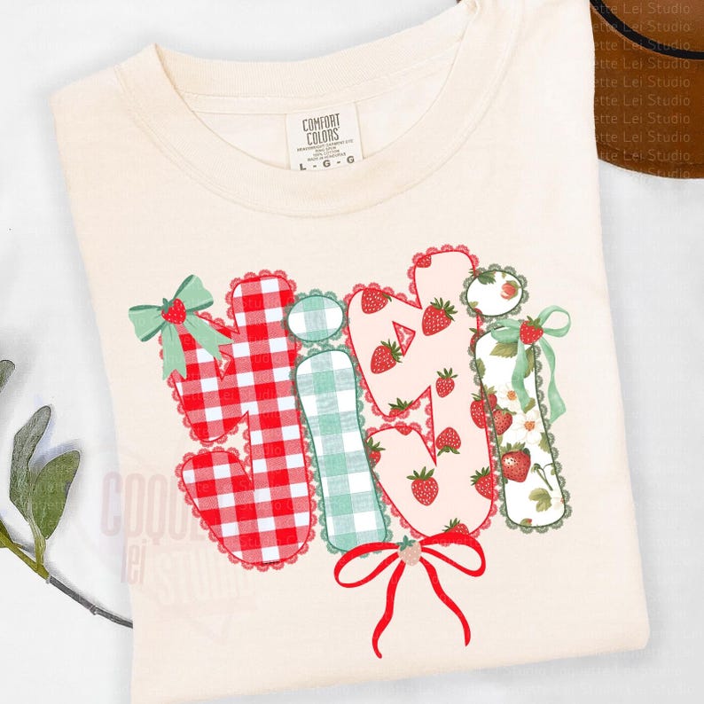 May include: Cream-colored Comfort Colors t-shirt with the word "Hi Ji Ji" in a playful, colorful design. The letters are decorated with gingham, strawberries, and bows. The shirt has a crew neck and short sleeves.