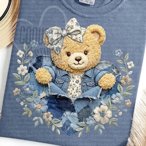 May include: A light blue sweatshirt featuring a teddy bear design. The bear is wearing a denim jacket and bow, with a heart-shaped denim patch. Floral embroidery in blue and cream surrounds the bear, creating a charming and whimsical aesthetic.