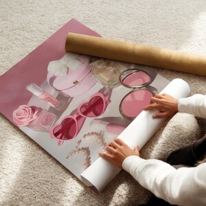 May include: A rolled-up print featuring a pink illustration of beauty items, including heart-shaped sunglasses, a perfume bottle, and a rose. A brown cardboard tube rests above the print. The print is on a white carpet.